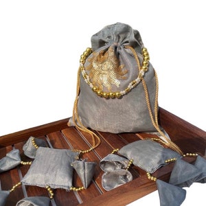 May include: A set of gray drawstring pouches with gold bead trim and a gold embroidered floral design. The pouches are displayed on a wooden tray.