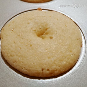 Puede incluir: Primer plano de una sola dona de color claro en un molde para donas. La dona est&aacute; completamente cocida y tiene un agujero en el centro.
