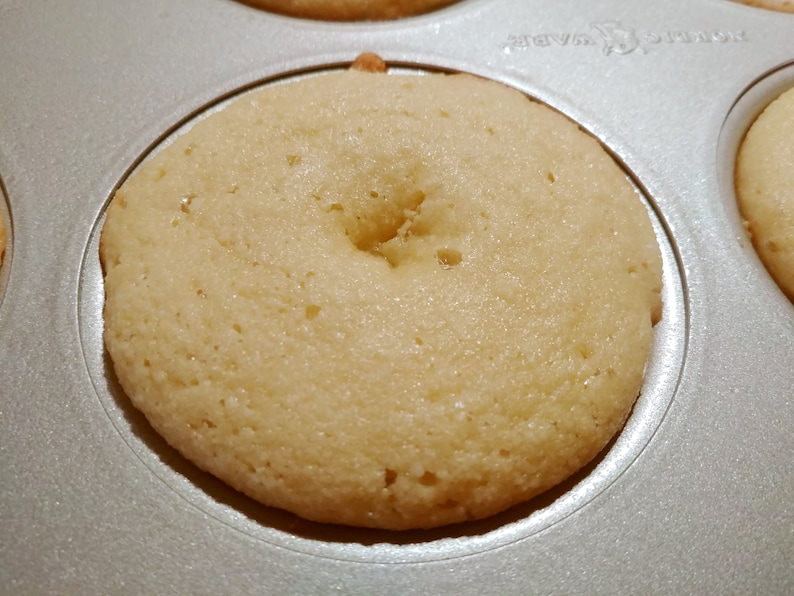 Puede incluir: Primer plano de una sola dona de color claro en un molde para donas. La dona est&aacute; completamente cocida y tiene un agujero en el centro.