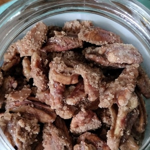 May include: A clear glass jar filled with candied pecan halves. The pecans are coated in a light brown sugar and have a crystallized appearance. The jar is sitting on a light blue surface.