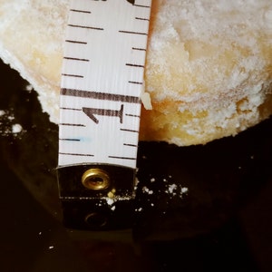 Puede incluir: Primer plano de una cinta m&eacute;trica blanca con marcas negras, midiendo una galleta redonda, blanca y en polvo. La cinta indica "1" y "2".
