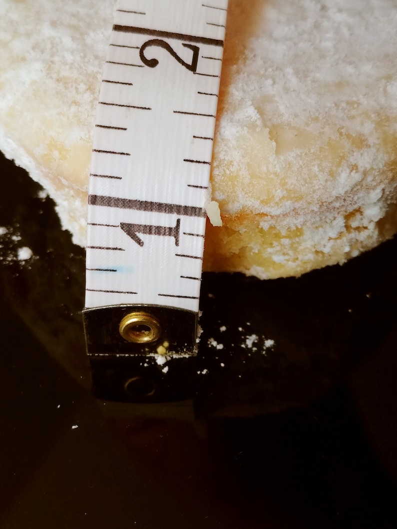 Puede incluir: Primer plano de una cinta m&eacute;trica blanca con marcas negras, midiendo una galleta redonda, blanca y en polvo. La cinta indica "1" y "2".