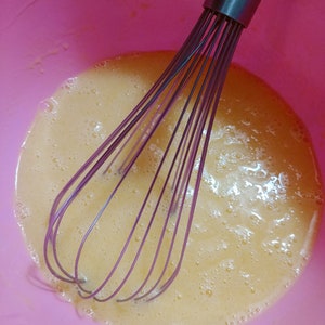 Puede incluir: Primer plano de un bol mezclador rosa con un batidor dentro. El bol est&aacute; lleno de un l&iacute;quido amarillo claro, probablemente yemas de huevo, que se ha batido hasta que est&eacute; espumoso.