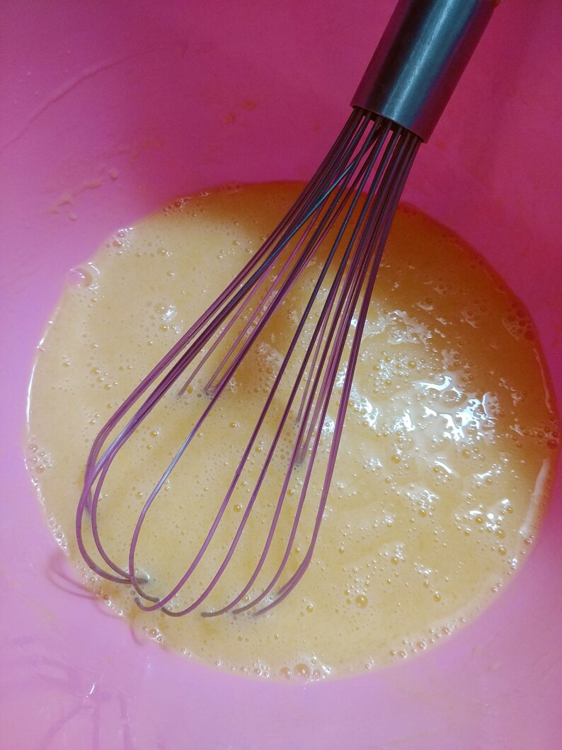 Puede incluir: Primer plano de un bol mezclador rosa con un batidor dentro. El bol est&aacute; lleno de un l&iacute;quido amarillo claro, probablemente yemas de huevo, que se ha batido hasta que est&eacute; espumoso.