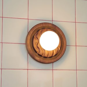 May include: A round, wooden wall sconce with a bright, white light. The sconce has a layered design with a carved, wavy pattern around the light. The sconce is mounted on a white tile wall with red grout lines.