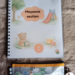 K&ouml;nnte beinhalten: Ein Spiralbuch mit dem Text "Moyenne section" und "4/5 ans" auf dem Cover, mit Illustrationen eines Spielplatzes, eines Teddyb&auml;ren und Bl&ouml;cken. Ein durchsichtiges Federm&auml;ppchen mit bunten Formen befindet sich darunter.