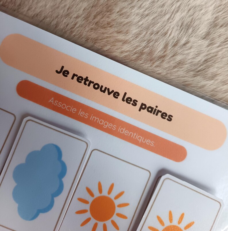 K&ouml;nnte beinhalten: Ein Kinderspiel mit dem Text "Je retrouve les paires" und "Associe les images identiques". Das Spiel zeigt Karten mit Illustrationen einer blauen Wolke und einer orangefarbenen Sonne. Der Hintergrund ist eine hellbraune, strukturierte Oberfl&auml;che.