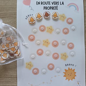 May include: A white chart with the French text "EN ROUTE VERS LA PROPRETÉ" and illustrations of stars, suns, and droplets. A small bag contains matching droplet-shaped tokens. The chart is designed for tracking progress.