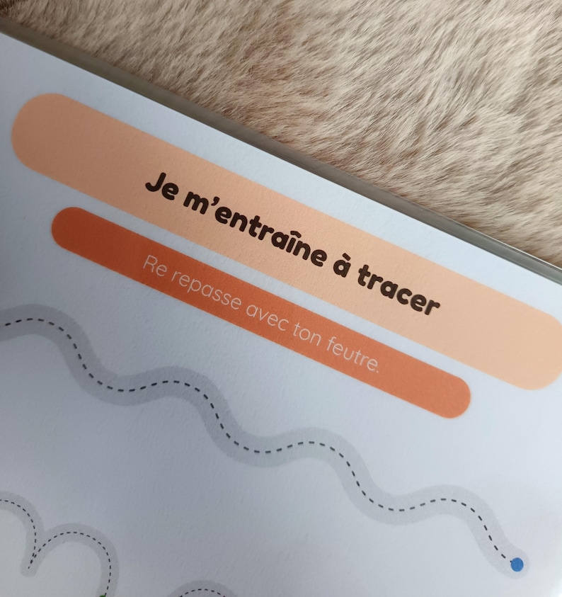 K&ouml;nnte beinhalten: Eine Seite aus einem Kinder-Aktivit&auml;tsbuch mit dem Text "Je m'entra&icirc;ne &agrave; tracer" und "Re repasse avec ton feutre". Die Seite hat einen hellblauen Hintergrund mit einer gepunkteten Linie zum Nachzeichnen. Zwei orangefarbene und pfirsichfarbene Rechtecke befinden sich oben.