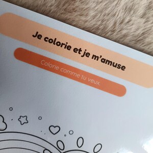 K&ouml;nnte beinhalten: Eine hellblaue Malbuchseite mit dem Text "Je colorie et je m'amuse" und "Colorie comme tu veux" in Schwarz. Die Seite hat eine Zeichnung eines Regenbogens, Herzen und Sterne.