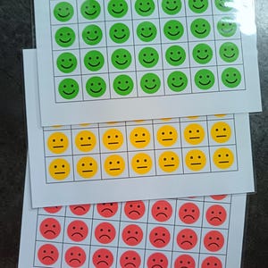 Puede incluir: Tres hojas de papel con una cuadrícula de 36 círculos cada una. Los círculos están llenos de caritas sonrientes de diferentes colores. La hoja superior tiene caritas sonrientes verdes, la hoja del medio tiene caritas sonrientes amarillas y la hoja inferior tiene caritas sonrientes rojas.