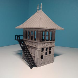May include: A grey model railway signal tower with a black metal staircase. The tower has a gabled roof and multiple windows.