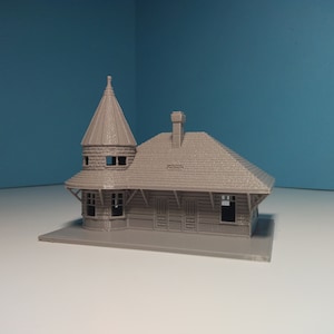 Puede incluir: Un modelo de estación de tren pequeña de color gris impreso en 3D con una torre y un techo. El modelo tiene un exterior detallado con ventanas y puertas.