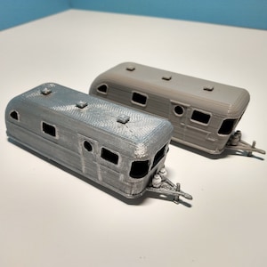 May include: Two miniature gray travel trailers. The trailers have rounded corners, small windows, and detailed hitches. The trailers are likely 3D printed and are designed to be used as toys or decorative items.