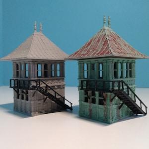 Puede incluir: Dos torres de señalización de tren en miniatura, una en gris y otra en verde con un techo rojo oxidado. Las torres tienen ventanas detalladas y una pequeña escalera.