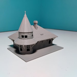 May include: A grey miniature model of a building with a conical tower, a chimney, and a sloped roof. The building has multiple windows and is set on a rectangular base. The model is likely for a train set or architectural display.
