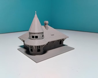 Op de afbeelding: Een grijs miniatuurmodel van een gebouw met een conische toren, een schoorsteen en een schuin dak. Het gebouw heeft meerdere ramen en staat op een rechthoekige basis. Het model is waarschijnlijk voor een modeltrein of architectuurdisplay.