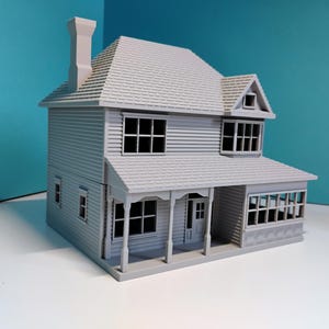 May include: A grey, two-storey model house with a gabled roof, chimney, and multiple windows. The house has a porch with columns and a front door. The siding is horizontal and the roof is shingled. The model is set against a teal background.