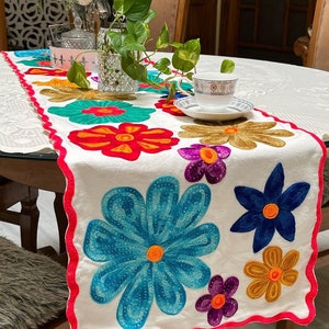 Puede incluir: Camino de mesa blanco con flores bordadas de colores, rojas, azules, amarillas y moradas. El camino de mesa tiene un borde festoneado con un ribete rosa brillante. Una taza y un platillo están colocados encima.