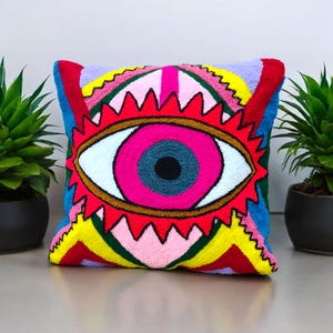 Puede incluir: Una almohada cuadrada colorida con un gran ojo bordado en el centro. El ojo está rodeado de un patrón geométrico rojo, amarillo y azul.