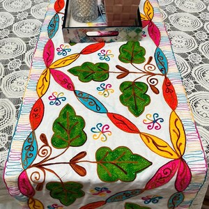 Puede incluir: Caminero de mesa blanco con hojas y espirales bordadas de colores. El camino de mesa tiene un borde rosa, amarillo, azul y verde.