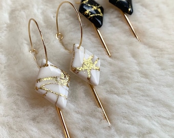 Puede incluir: Pendientes de aro dorados con colgantes geométricos. Los pendientes presentan aros dorados y colgantes en forma de diamante. Los colgantes son blancos con vetas doradas y negros con vetas doradas. Una punta dorada se extiende desde la parte inferior de cada colgante.