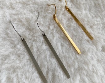 Puede incluir: Tres pares de pendientes minimalistas de barra en tonos plateados y dorados. Los pendientes se muestran sobre una superficie suave, blanca y texturizada. Los pendientes tienen un diseño lineal simple, con cierre de gancho.