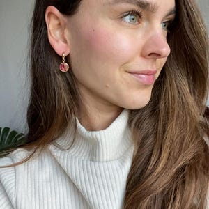May include: Gold hoop earrings featuring a small, round, pink and red floral design. The earrings are worn by a person with long, brown hair and a white turtleneck jumper. The earrings are the main focus.