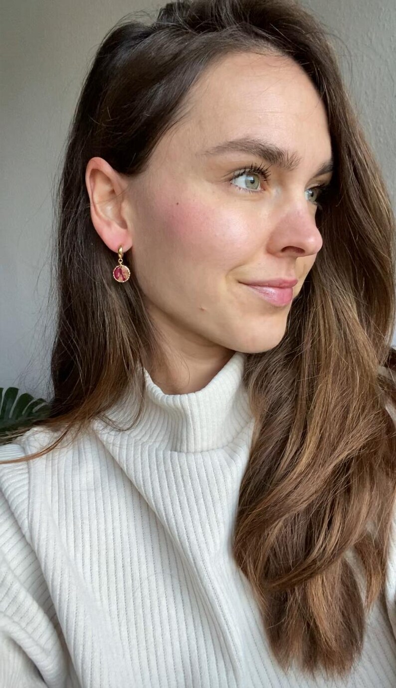 May include: Gold hoop earrings featuring a small, round, pink and red floral design. The earrings are worn by a person with long, brown hair and a white turtleneck jumper. The earrings are the main focus.