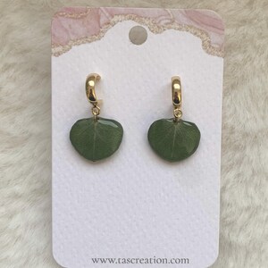 Pode incluir: Brincos de argola dourados com pingentes verdes em forma de folha. S&atilde;o exibidos em um cart&atilde;o branco com uma borda decorativa rosa e dourada. O endere&ccedil;o do site www.tascreation.com est&aacute; impresso na parte inferior.
