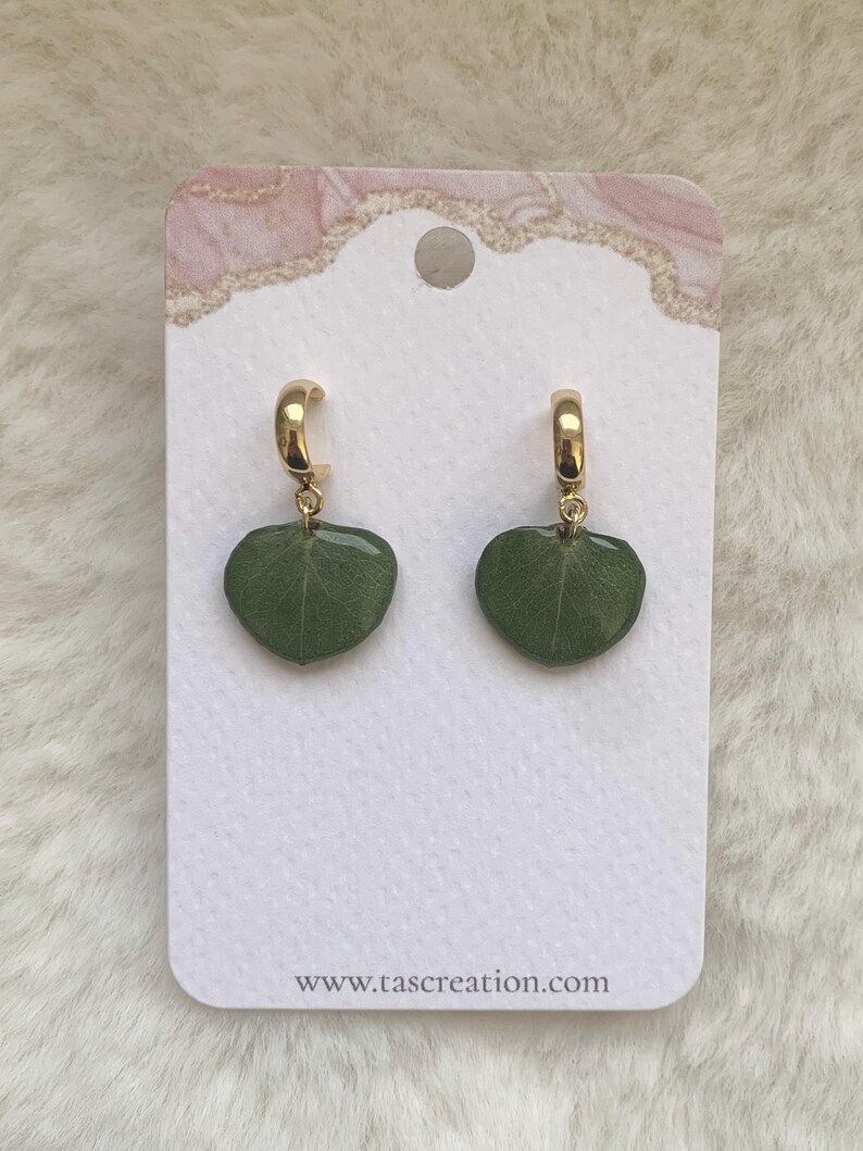Pode incluir: Brincos de argola dourados com pingentes verdes em forma de folha. S&atilde;o exibidos em um cart&atilde;o branco com uma borda decorativa rosa e dourada. O endere&ccedil;o do site www.tascreation.com est&aacute; impresso na parte inferior.