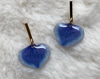 Puede incluir: Un par de pendientes colgantes con colgantes en forma de corazón hechos de pétalos de hortensia azules preservados e incrustados en resina transparente brillante. Cada pétalo está unido a una fina barra metálica dorada vertical.
