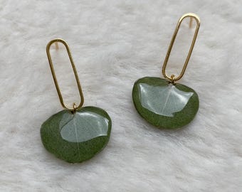 Puede incluir: Un par de pendientes colgantes con aros ovalados alargados de tono dorado unidos a pequeñas hojas verdes redondeadas y brillantes. Las hojas tienen un acabado liso y reflectante que resalta las venas naturales y la textura del material.