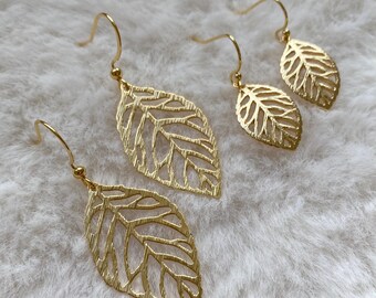 Puede incluir: Dos pares de pendientes dorados con forma de hoja. Los pendientes presentan un diseño calado que muestra las venas de las hojas. Se exhiben sobre una superficie suave y blanca. Ligeros, son perfectos para el uso diario.