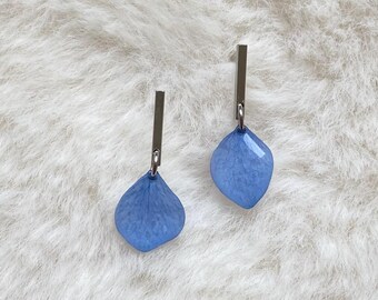 May include: A pair of blue petal-shaped earrings with silver rectangular posts. The earrings are a light blue colour and have a faceted texture. The earrings are displayed on a white, fluffy surface.