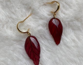 Puede incluir: Un par de pendientes de aro dorados con colgantes en forma de hoja de color rojo oscuro. Los colgantes tienen un acabado brillante y translúcido con detalles de venas oscuras, suspendidos de aros de metal dorado pulido con cierre de poste.