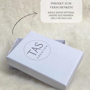 May include: A white gift box with the text "TAS CREATION" in a circular logo. The box is open, revealing its interior. Text on the image reads "PERFEKT ZUM VERSCHENKEN! W&Auml;HLE DAF&Uuml;R OPTIONAL UNSERE GESCHENKBOX (9x6.5 cm) DAZU AUS."