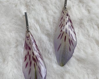 May include: A pair of drop earrings featuring preserved, light purple flower petals with dark purple speckles. Each petal is encased in a clear, glossy resin coating and attached to a slender, polished silver-tone metal bar post finding.