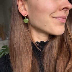 Pode incluir: Um brinco de argola dourado com um pendente de pedra verde em forma de cora&ccedil;&atilde;o. A pessoa tem cabelo castanho comprido e usa um top preto. Joia de moda.