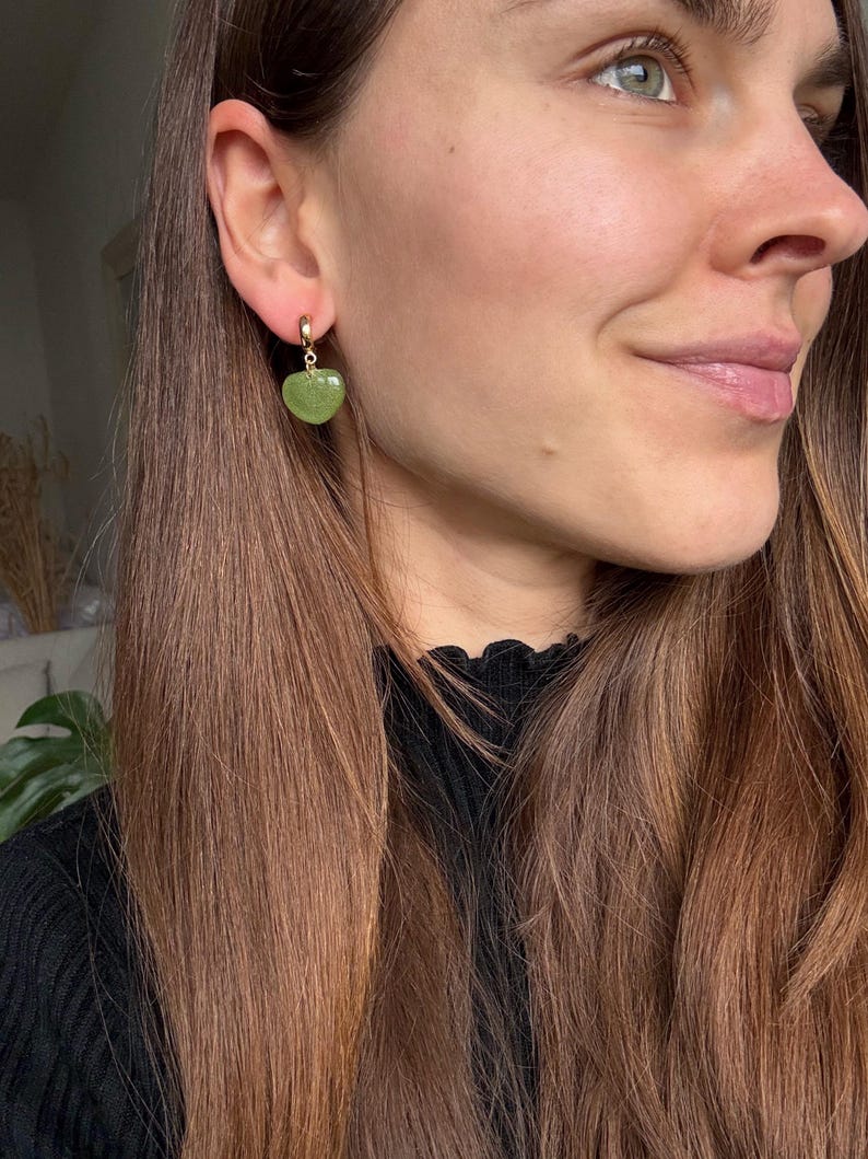 Pode incluir: Um brinco de argola dourado com um pendente de pedra verde em forma de cora&ccedil;&atilde;o. A pessoa tem cabelo castanho comprido e usa um top preto. Joia de moda.