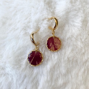 May include: A pair of gold-coloured hoop earrings. Each earring has a circular, burgundy-coloured pendant with a gold-coloured trim. The earrings are displayed on a white, fluffy surface.