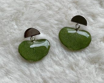 Puede incluir: Un par de pendientes con una pieza superior plateada en forma de semicírculo conectada a una hoja verde con forma de corazón, encapsulada en resina transparente y brillante. La textura natural de la hoja es visible a través de la resina.