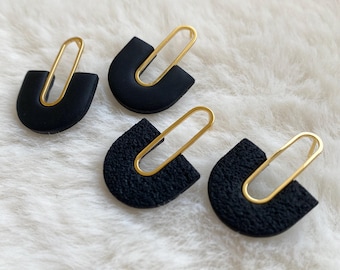 Puede incluir: Cuatro pendientes negros en forma de U con una superficie texturizada, cada uno con un diseño de clip de papel dorado alargado. Los pendientes se muestran sobre una superficie blanca y esponjosa, creando un contraste.