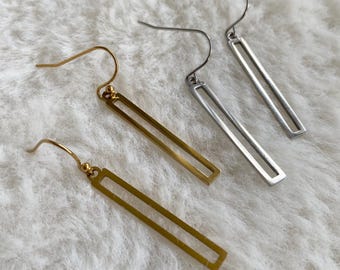 Puede incluir: Primer plano de cuatro pares de pendientes. Dos pares son dorados y dos plateados. Cada pendiente presenta un diseño de barra rectangular suspendido de un alambre curvo. Los pendientes se muestran sobre una superficie blanca y esponjosa.