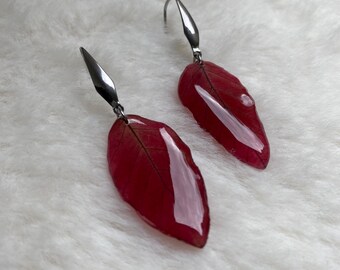 May include: A pair of drop earrings featuring preserved, deep red leaves coated in a glossy, clear resin finish. Each leaf is attached to a polished silver-tone metal hook finding that tapers to a point, creating a sleek and reflective appearance.