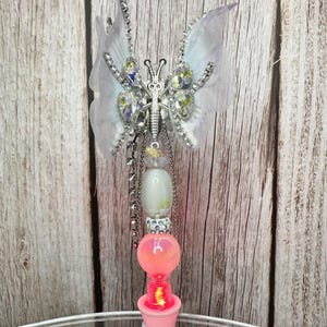 Puede incluir: Una varita decorativa de mariposa con un cuerpo y alas de metal plateado. Las alas son blancas con cristales transparentes y un contorno plateado. La varita tiene una base rosa y blanca con un orbe rosa brillante y una cadena de cuentas plateadas.