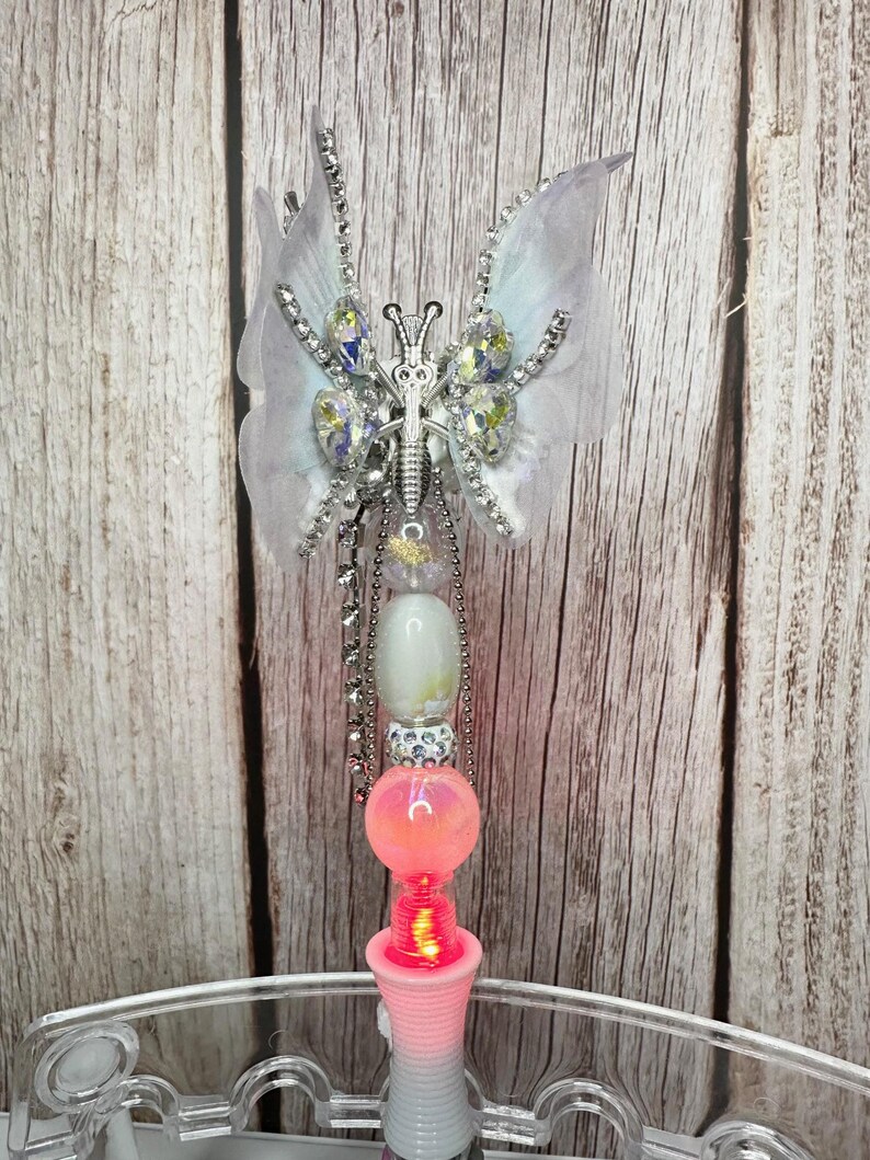 Puede incluir: Una varita decorativa de mariposa con un cuerpo y alas de metal plateado. Las alas son blancas con cristales transparentes y un contorno plateado. La varita tiene una base rosa y blanca con un orbe rosa brillante y una cadena de cuentas plateadas.