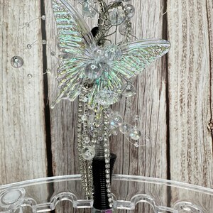 Puede incluir: Un tap&oacute;n de botella decorativo con un dise&ntilde;o de mariposa transparente e iridiscente. La mariposa est&aacute; adornada con cuentas transparentes y detalles plateados. El tap&oacute;n tiene un cuerpo negro y transparente con un acento rosa.