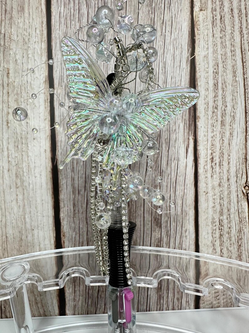 Puede incluir: Un tap&oacute;n de botella decorativo con un dise&ntilde;o de mariposa transparente e iridiscente. La mariposa est&aacute; adornada con cuentas transparentes y detalles plateados. El tap&oacute;n tiene un cuerpo negro y transparente con un acento rosa.