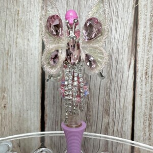 Puede incluir: Un bol&iacute;grafo decorativo con dise&ntilde;o de mariposa. La mariposa tiene alas de encaje blanco adornadas con gemas rosas. El cuerpo del bol&iacute;grafo es lavanda y el cuerpo de la mariposa est&aacute; adornado con pedrer&iacute;a.